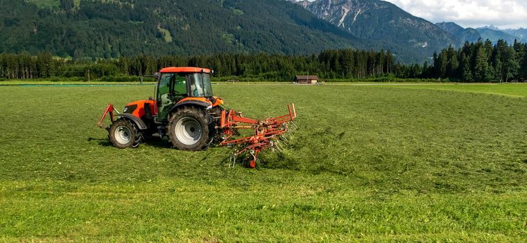 Agricultural machinery, a tractor collecting grass in a field against a blue sky. Season harvesting, grass, agricultural land.