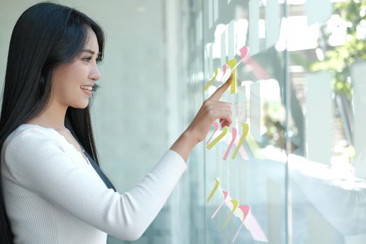 businesswoman woman thinking planning with adhesive notes on glass wall at workplace. Sticky note paper reminder schedule for creative idea & business brainstorming