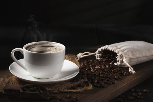 White cup of fresh hot coffee and coffee beans on wooden board on table.