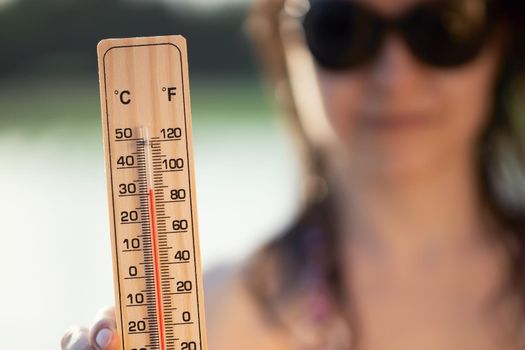 Beautiful young woman in sunglasses with a thermometer in her hand on the background of the lake. Summer vacation and heat concept, selective focus.