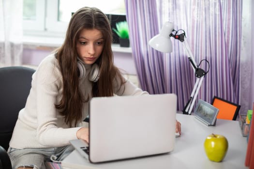 A young brunette with long hair sits at home at a desk next to a window watching a school lecture. A pretty girl focused on learning.