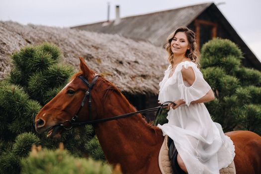 A woman in a white sundress riding a horse near a farm.