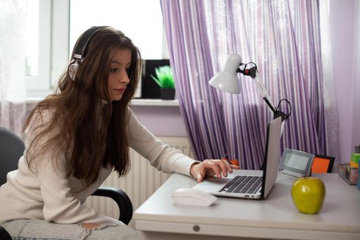 A young female student is taking schoolwork via the internet. An attractive brunette with long hair sits at a desk near a window.