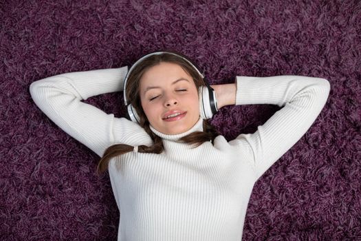 Happy brunette with eyes closed listens to music on wireless headphones while lying on purple carpet. Musical relaxation.