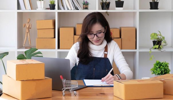 Young beautiful happy asian business woman owner of SME online using laptop receive order from customer with parcel box packaging at her startup home office, online business seller and delivery.