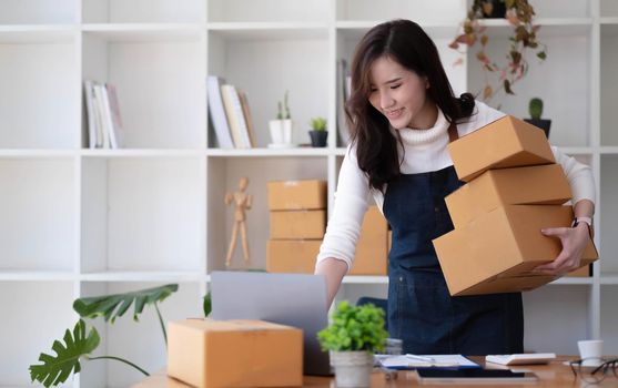 Young beautiful happy asian business woman owner of SME online using laptop receive order from customer with parcel box packaging at her startup home office, online business seller and delivery.