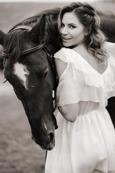 A girl in a white sundress stands next to a brown horse in a field in summer.black and white photo.