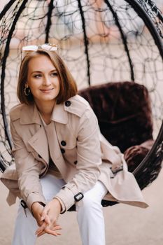 A happy stylish girl In a gray coat is sitting outside in an armchair.