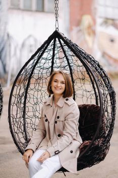 A happy stylish girl In a gray coat is sitting outside in an armchair.