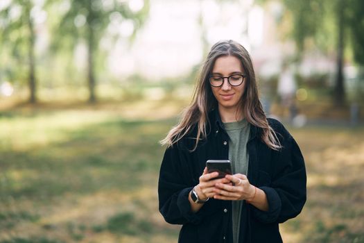 Young woman with glasses reading a message on her phone while standing in the park. High quality photo