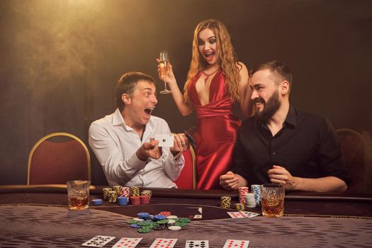 Two handsome friends and gorgeous girl are playing poker at casino. They are very happy about their win, rejoicing , laughing and having a good time while posing at the table against a yellow backlight on smoke background. Cards, chips, money, gambling, entertainment concept.