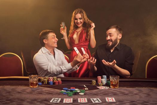 Two athletic friends and charming maiden are playing poker at casino. They are rejoicing their win and showing the winning combination to each other while posing at the table against a yellow backlight on smoke background. Cards, chips, money, gambling, entertainment concept.