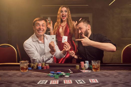 Two athletic friends and charming girl are playing poker at casino. They are rejoicing their win, smiling and showing the winning combination while posing at the table against a yellow backlight on smoke background. Cards, chips, money, gambling, entertainment concept.