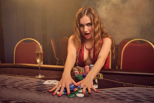 Gorgeous woman with a long curly hair and perfect make-up, dressed in a sexy red dress. She is sitting at a gambling table, holding some chips and looking at the camera. Poker concept on a dark smoke background in a ray of a yellow spotlight. Casino.