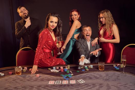 Joyful partners are playing poker at casino. They are celebrating their win, smiling and looking vey excited while posing at the table against a dark background. Cards, chips, money, alcohol, gambling, entertainment concept.