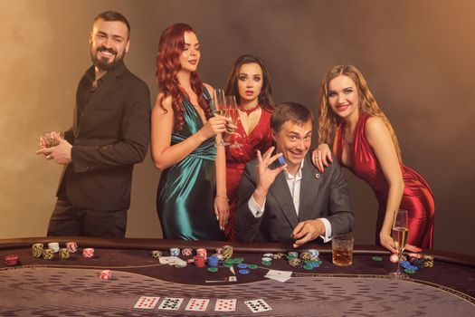 Funny classmates are playing poker at casino. They are celebrating their win, smiling and looking vey excited while posing at the table against a dark smoke background in a ray of a spotlight. Cards, chips, money, alcohol, gambling, entertainment concept.