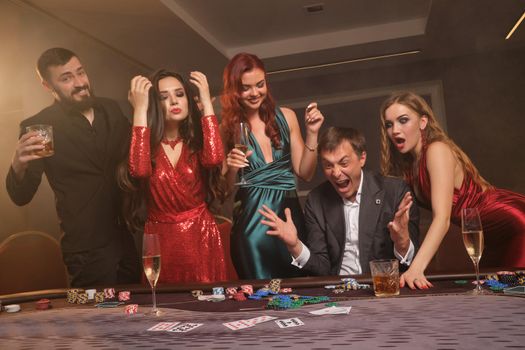 Enthusiastic friends are playing poker at casino. They are celebrating their win, smiling and looking vey excited while posing at the table against a dark smoke background. Cards, chips, money, alcohol, gambling, entertainment concept.