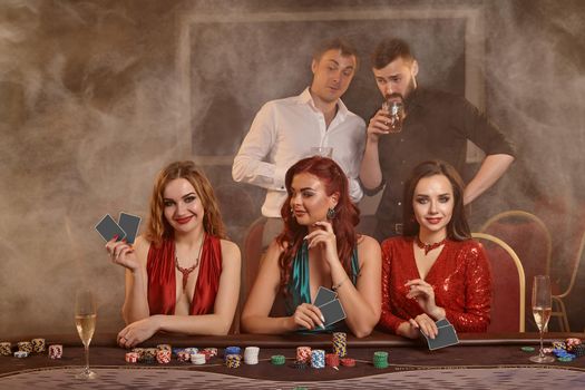 Concentrated companions are playing poker at casino. Golden youth are making bets waiting for a big win while posing at the table against a dark smoke background. Cards, chips, money, fortune, alcohol, gambling, entertainment concept.