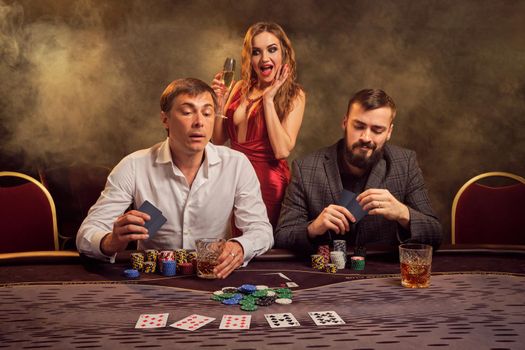 Two good-looking friends and sexy girl are playing poker at casino. They are making bets waiting for a big win, smiling and having a good time while posing at the table against a yellow backlight on smoke background. Cards, chips, money, gambling, entertainment concept.