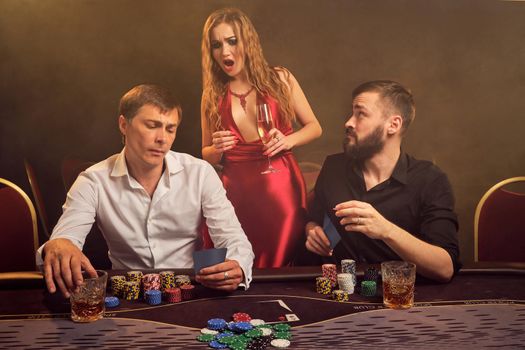Two old friends and attractive girl are playing poker at casino. They are making bets waiting for a big win, but looking a little bit shocked while posing at the table against a yellow backlight on smoke background. Cards, chips, money, gambling, entertainment concept.