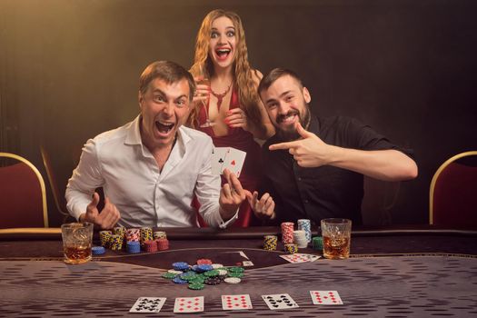 Two athletic friends and charming woman are playing poker at casino. They are rejoicing their win, smiling and looking very happy while posing at the table against a yellow backlight on smoke background. Cards, chips, money, gambling, entertainment concept.