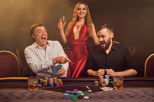 Two handsome friends and gorgeous lady are playing poker at casino. They are happy about their win, rejoicing and smiling while posing at the table against a yellow backlight on smoke background. Cards, chips, money, gambling, entertainment concept.
