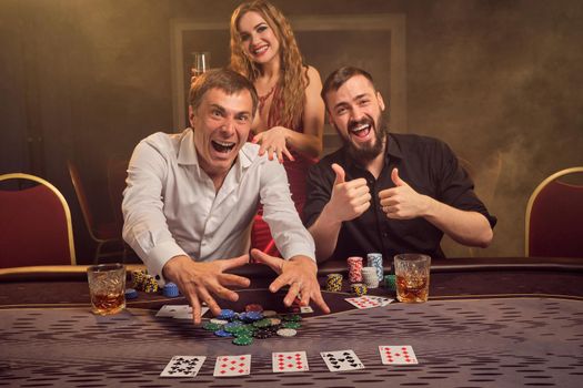 Two wealthy friends and beautiful lady are playing poker at casino. They are rejoicing their win, smiling and having a good time while posing at the table against a yellow backlight on smoke background. Cards, chips, money, gambling, entertainment concept.