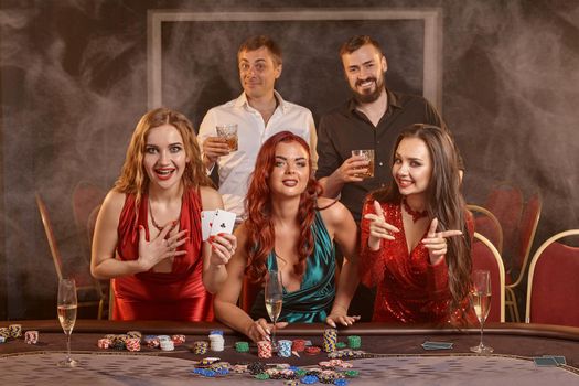 Fortunate colleagues are playing poker at casino. They are celebrating their win, smiling and posing at the table against a dark smoke background. Cards, chips, money, alcohol, gambling, entertainment concept.