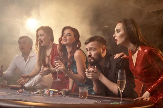Cheerful friends are playing poker at casino. Youth are making bets waiting for a big win while posing at the table against a white spotlight on a dark smoke background. Cards, chips, money, alcohol, gambling, entertainment concept.