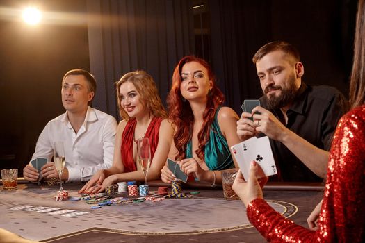 Enthusiastic companions are playing poker at casino. Youth are making bets waiting for a big win while posing at the table against a white spotlight on a dark smoke background. Cards, chips, money, alcohol, gambling, entertainment concept.