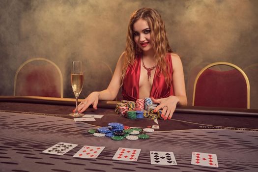 Attractive girl with a long curly hair and perfect make-up, dressed in a sexy red dress. She is sitting at a gambling table and smiling. Poker concept on a dark smoke background in a ray of a yellow spotlight. Casino.