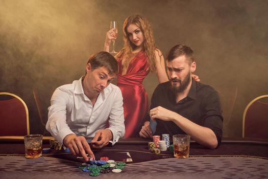 Two handsome friends and gorgeous maiden are playing poker at casino. They are making bets waiting for a big win and having a good time while posing at the table against a yellow backlight on smoke background. Cards, chips, money, gambling, entertainment concept.