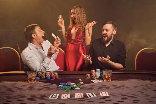 Two handsome friends and gorgeous woman are playing poker at casino. They are happy about their win, rejoicing and having a good time while posing at the table against a yellow backlight on smoke background. Cards, chips, money, gambling, entertainment concept.