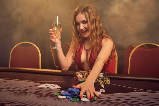 Alluring woman with a long curly hair and perfect make-up, dressed in a sexy red dress. She is sitting at a gambling table, holding some chips and champagne. Poker concept on a dark smoke background in a ray of a yellow spotlight. Casino.