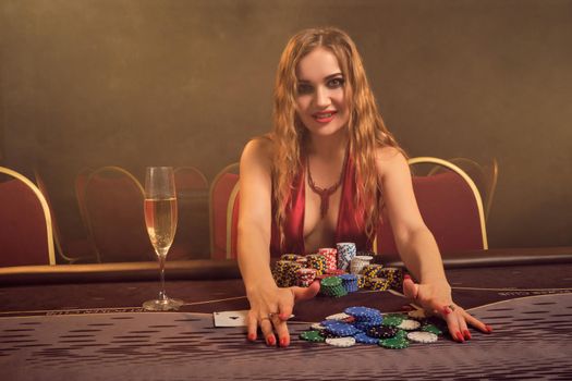 Gorgeous maiden with a long curly hair and perfect make-up, dressed in a sexy red dress. She is sitting at a gambling table, holding some chips and smiling. Poker concept on a dark smoke background in a ray of a yellow spotlight. Casino.