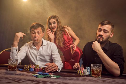 Two old friends and attractive woman are playing poker at casino. They are making bets waiting for a big win while posing at the table against a yellow backlight on smoke background. Cards, chips, money, gambling, entertainment concept.
