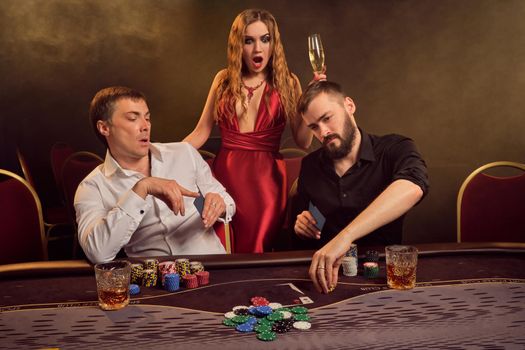 Two old friends and attractive maiden are playing poker at casino. They are making bets waiting for a big win and looking wondered while posing at the table against a yellow backlight on smoke background. Cards, chips, money, gambling, entertainment concept.