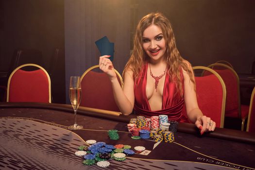 Beautiful woman with a long curly hair and perfect make-up, dressed in a sexy red dress. She is sitting at a gambling table with two playing cards in her hands and smiling. Poker concept on a dark smoke background in a ray of a yellow spotlight. Casino.