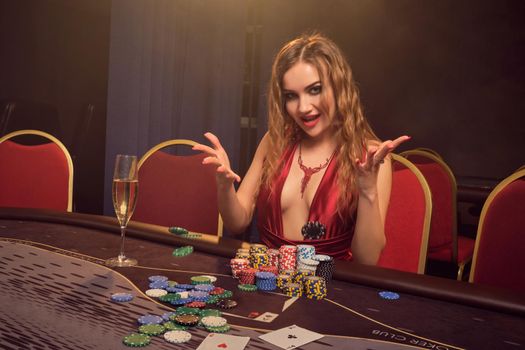 Pretty female with a long curly hair and perfect make-up, dressed in a sexy red dress. She is rejoicing her win while sitting at a gambling table and smiling. Poker concept on a dark smoke background in a ray of a yellow spotlight. Casino.