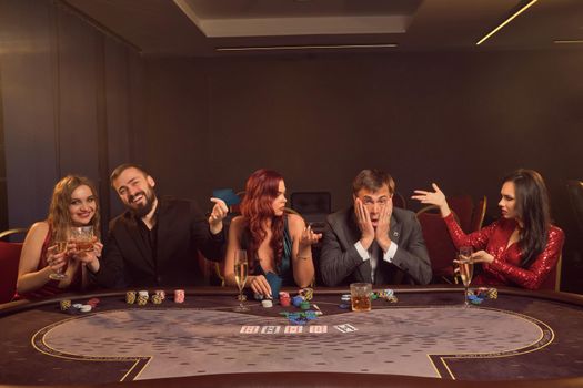 Merry buddies are playing poker at casino. They are making bets waiting for a big win while posing at the table against a dark smoke background in a ray of a spotlight. Cards, chips, money, alcohol, gambling, entertainment concept.