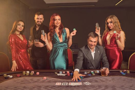 Happy partners are playing poker at casino. They are celebrating their win, smiling and looking vey excited while posing at the table against a dark smoke background in a ray of a spotlight. Cards, chips, money, alcohol, gambling, entertainment concept.