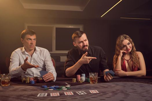 Two wealthy friends and beautiful girl are playing poker at casino. They are making bets waiting for a big win, looking at someone and having a good time while posing sitting at the table against a yellow backlight on dark background. Cards, chips, money, gambling, entertainment concept.