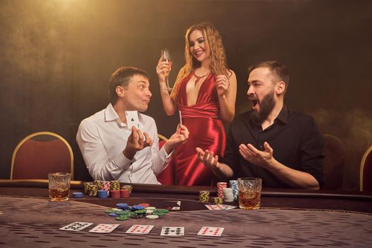 Two handsome friends and gorgeous female are playing poker at casino. They are happy about their win, rejoicing and smiling while posing at the table against a yellow backlight on smoke background. Cards, chips, money, gambling, entertainment concept.