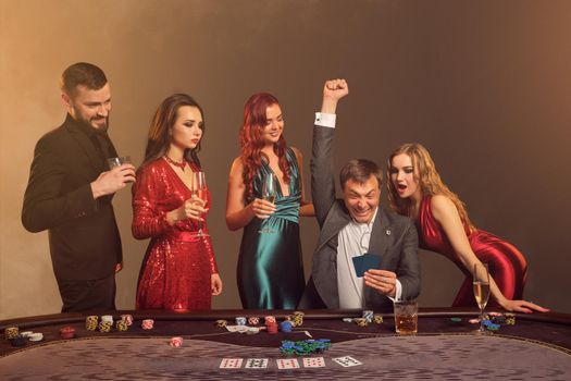 Funny buddies are playing poker at casino. They are celebrating their win, smiling and looking vey excited while posing at the table against a dark smoke background in a ray of a spotlight. Cards, chips, money, alcohol, gambling, entertainment concept.