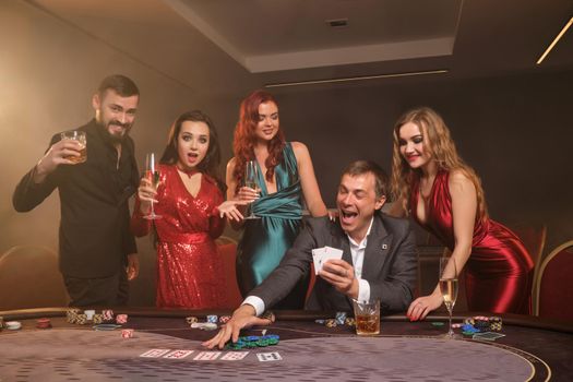 Enthusiastic classmates are playing poker at casino. They are celebrating their win, smiling and looking vey excited while posing at the table against a dark smoke background in a ray of a spotlight. Cards, chips, money, alcohol, gambling, entertainment concept.