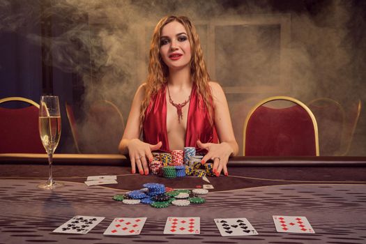 Attractive lady with a long curly hair and perfect make-up, dressed in a sexy red dress. She is sitting at a gambling table, holding some gambling chips and looking at the camera. Poker concept on a dark smoke background in a ray of a yellow spotlight. Casino.