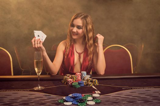 Alluring maiden with a long curly hair and perfect make-up, dressed in a sexy red dress. She is sitting at a gambling table with two playing cards in her hands and looking at them. Poker concept on a dark smoke background in a ray of a yellow spotlight. Casino.