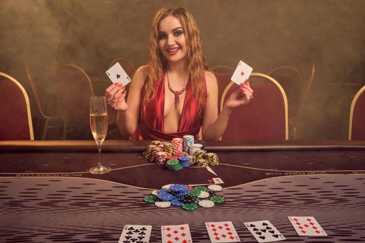 Attractive female with a long curly hair and perfect make-up, dressed in a sexy red dress. She is sitting at a gambling table with two playing cards in her hands and smiling. Poker concept on a dark smoke background in a ray of a yellow spotlight. Casino.