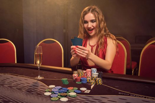 Beautiful girl with a long curly hair and perfect make-up, dressed in a sexy red dress. She is sitting at a gambling table with two playing cards in her hands and looking at it. Poker concept on a dark smoke background in a ray of a yellow spotlight. Casino.