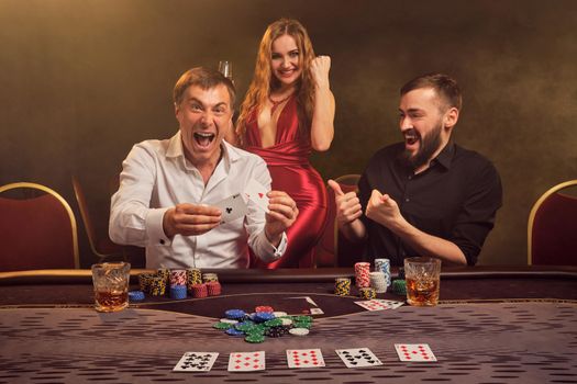 Two athletic friends and charming lady are playing poker at casino. They are rejoicing their win and showing two aces while posing at the table against a yellow backlight on smoke background. Cards, chips, money, gambling, entertainment concept.